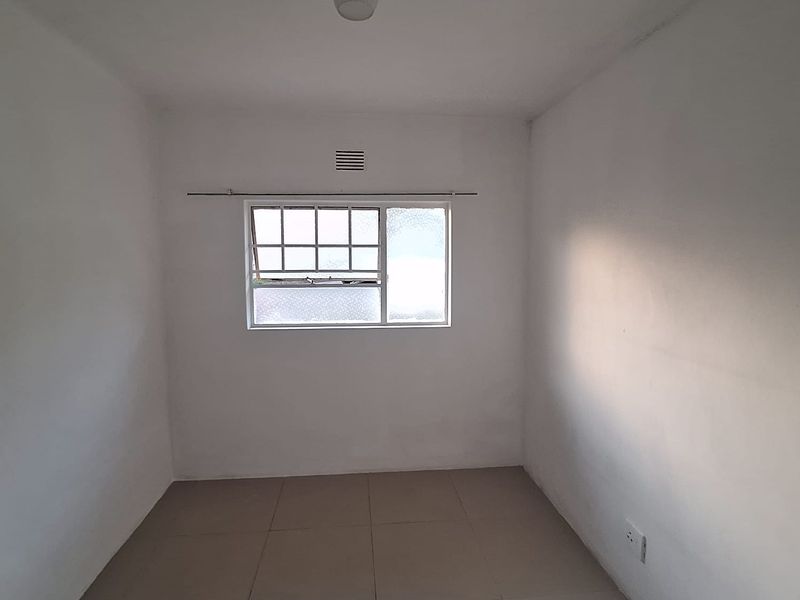 An empty, neutrally painted bedroom featuring tiled flooring, a window with security bars, and a central ceiling light fixture.