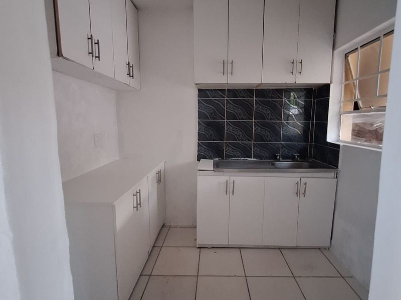 A compact, empty kitchen featuring white built-in cabinetry, a stainless steel sink, dark tiled backsplash, and tiled flooring.