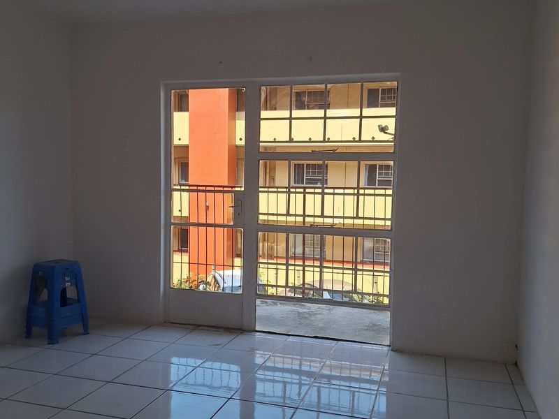 An empty living room featuring white tiled flooring, a ceiling light fixture, and a glass sliding door with a security gate leading to a balcony.