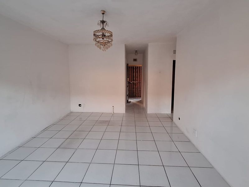 An empty, open-plan living area featuring white tiled flooring and a central ceiling light fixture, leading to a hallway entrance.