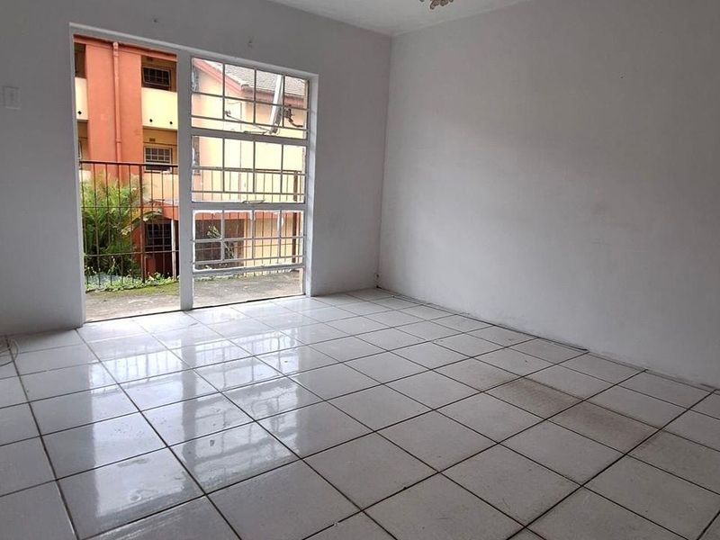 An empty living room featuring white tiled flooring and a large window with security bars, offering a blank canvas for potential interior design.