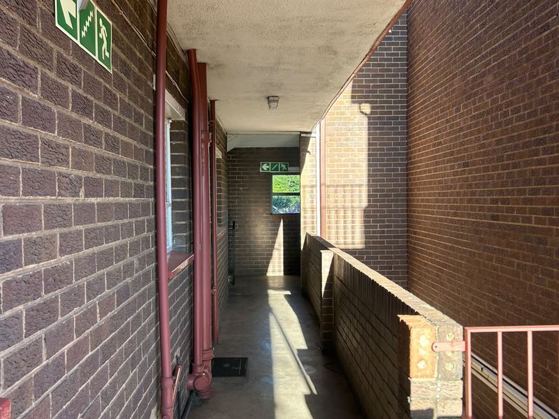 An exterior apartment building walkway featuring exposed brick walls, concrete flooring, and metal safety railings.