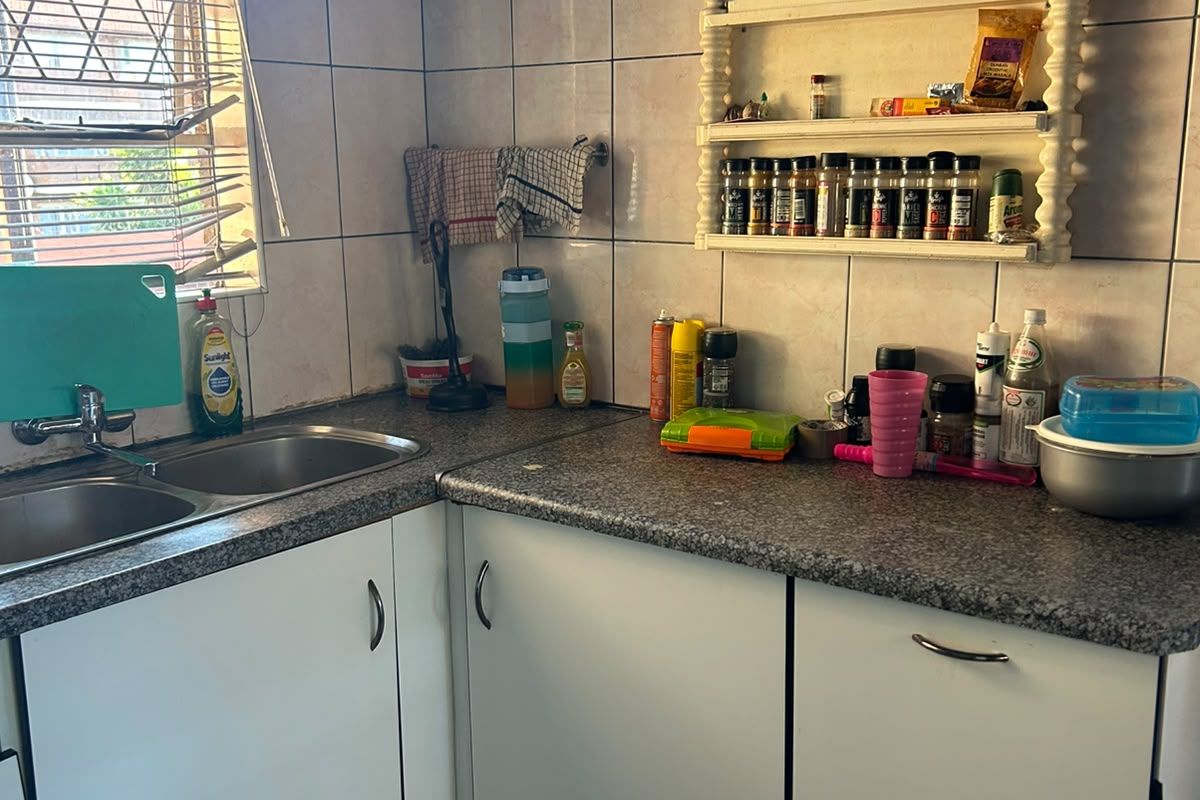 A compact kitchen featuring a double basin sink, laminate countertops, tiled backsplash, and built-in cabinetry with an overhead geyser unit.