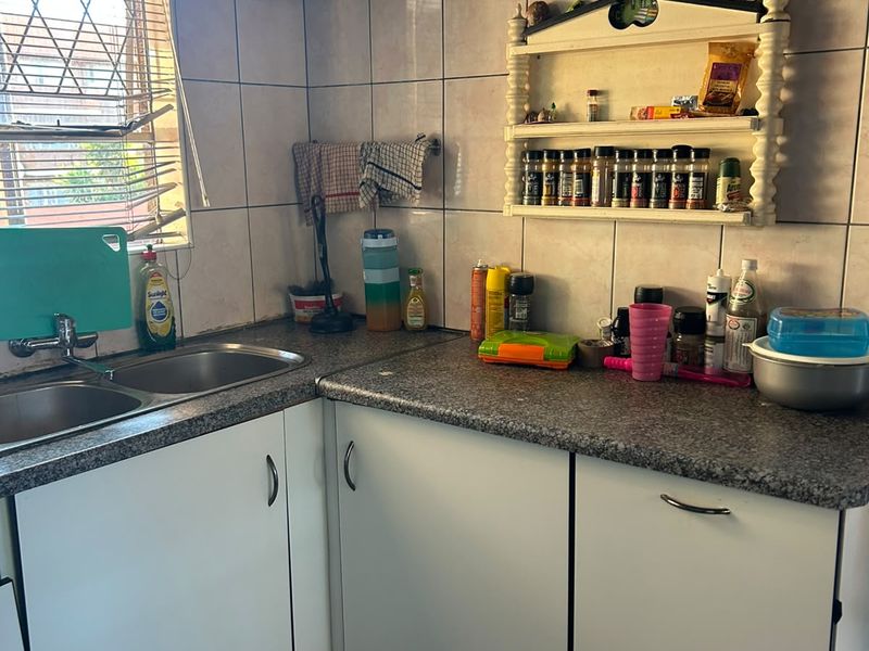 A compact kitchen featuring a double basin sink, laminate countertops, tiled backsplash, and built-in cabinetry with an overhead geyser unit.