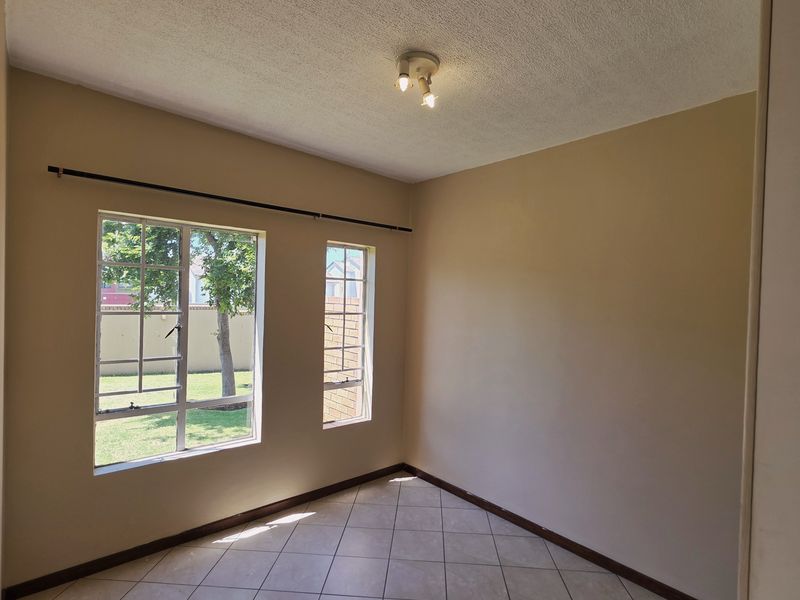 A bright, empty bedroom featuring neutral wall paint, tiled flooring, and large windows providing natural light and garden views.