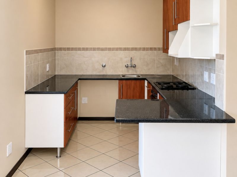 A clean, empty kitchen featuring granite countertops, built-in hob, tiled backsplash, and a mix of white and wood-finish cabinetry with tiled flooring.