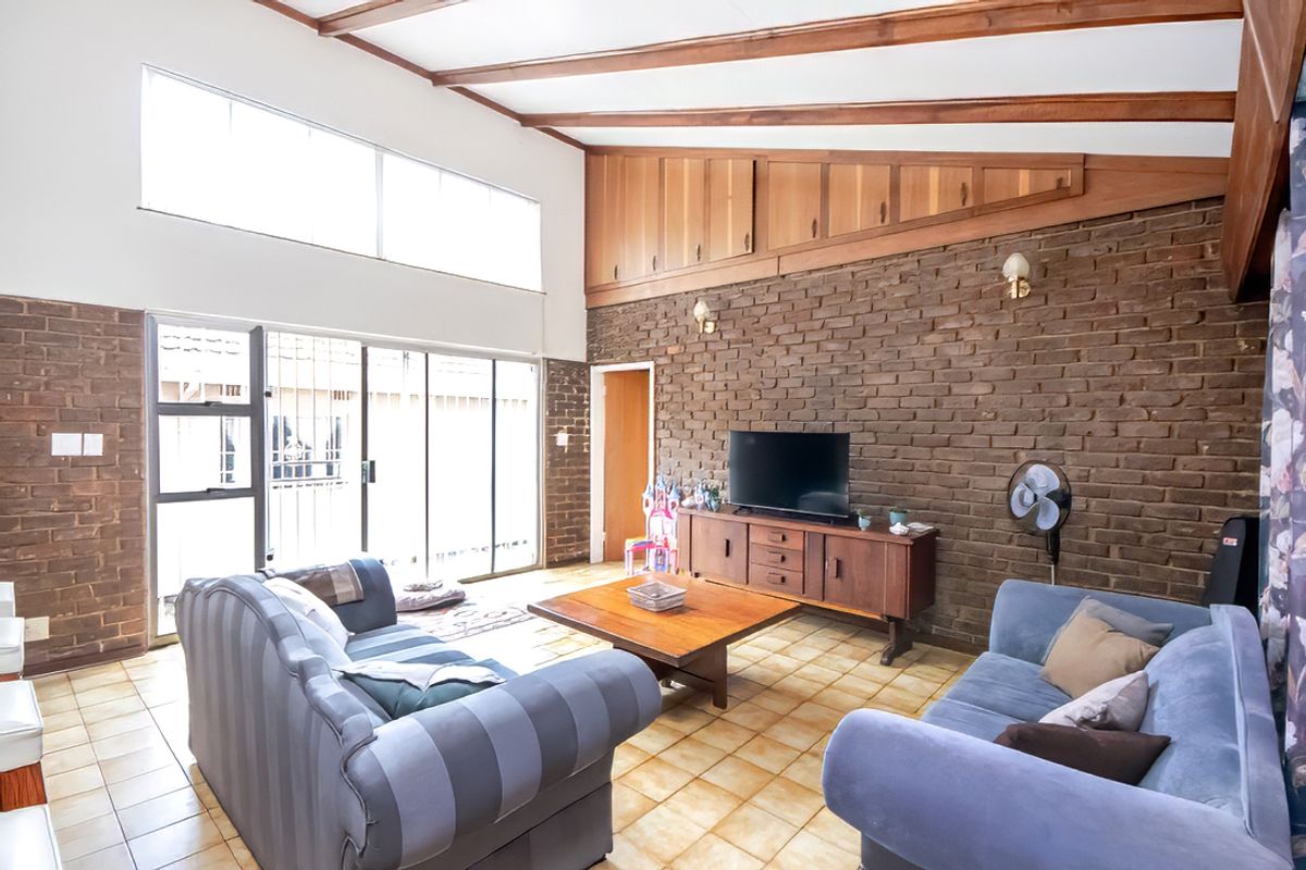 Spacious living room featuring a high vaulted ceiling with exposed timber beams, a prominent face-brick feature wall, and large windows providing ample natural light.