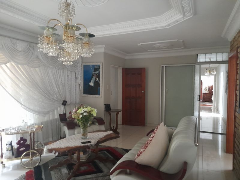 A spacious living room featuring decorative ceiling cornices, a crystal chandelier, and tiled flooring, leading into a connecting hallway.