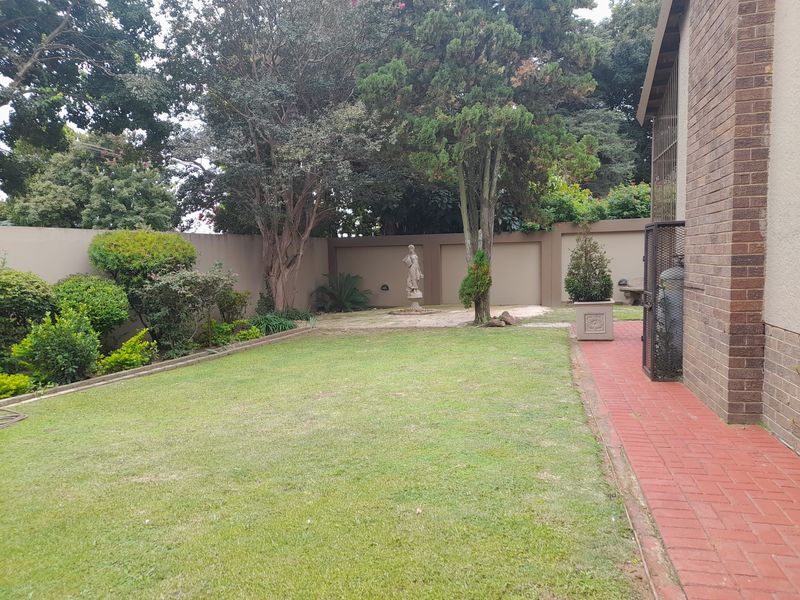 A private rear garden featuring a manicured lawn, established trees, perimeter boundary walls, and a red brick paved walkway along the side of the house.