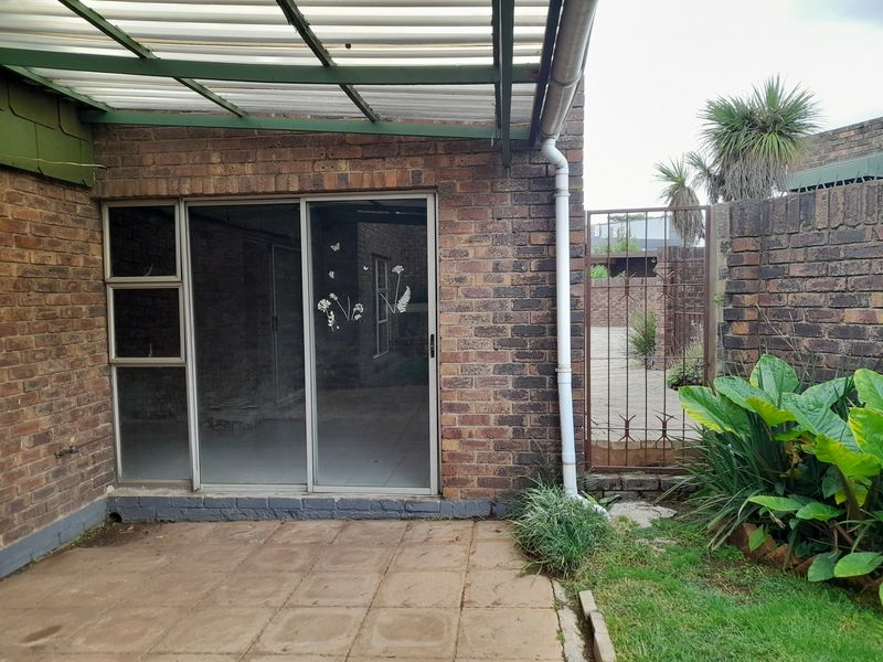A rear patio area featuring a covered roof structure, sliding glass doors, and a brick exterior wall with an adjacent metal gate.