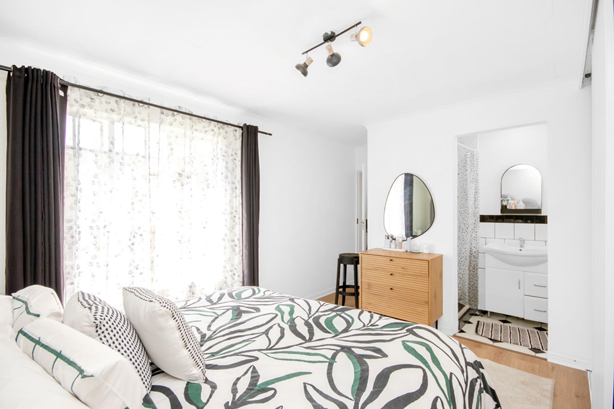 A bright bedroom featuring wood-look flooring, a modern track light fixture, and an en-suite bathroom with a vanity and tiled splashback.