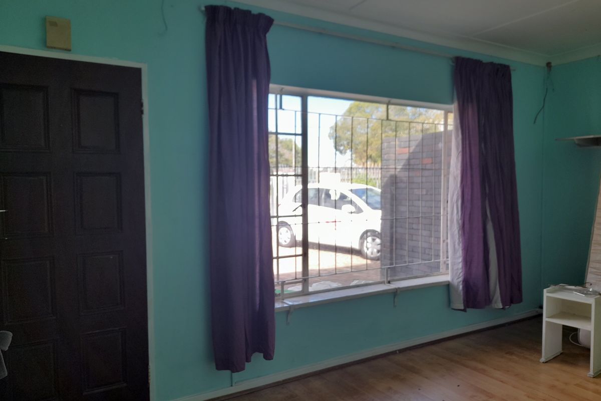 A living room featuring light blue walls, timber-look flooring, and a large window with security bars, providing natural light and street views.
