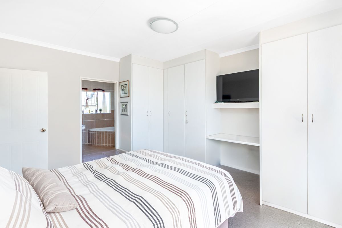 A bright bedroom featuring built-in cupboards, a dedicated desk area, and an en-suite bathroom with a tiled bathtub.