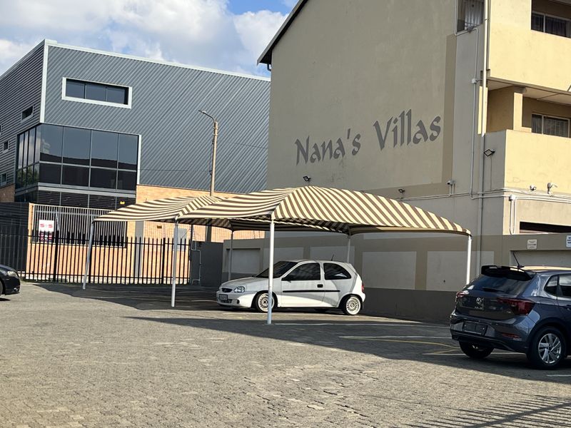Exterior view of Nana's Villas apartment complex featuring a paved parking area with a striped shade cloth carport structure.
