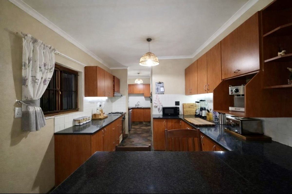 A functional kitchen featuring wooden cabinetry, dark granite countertops, tiled splashbacks, and integrated lighting, with a connected scullery area.