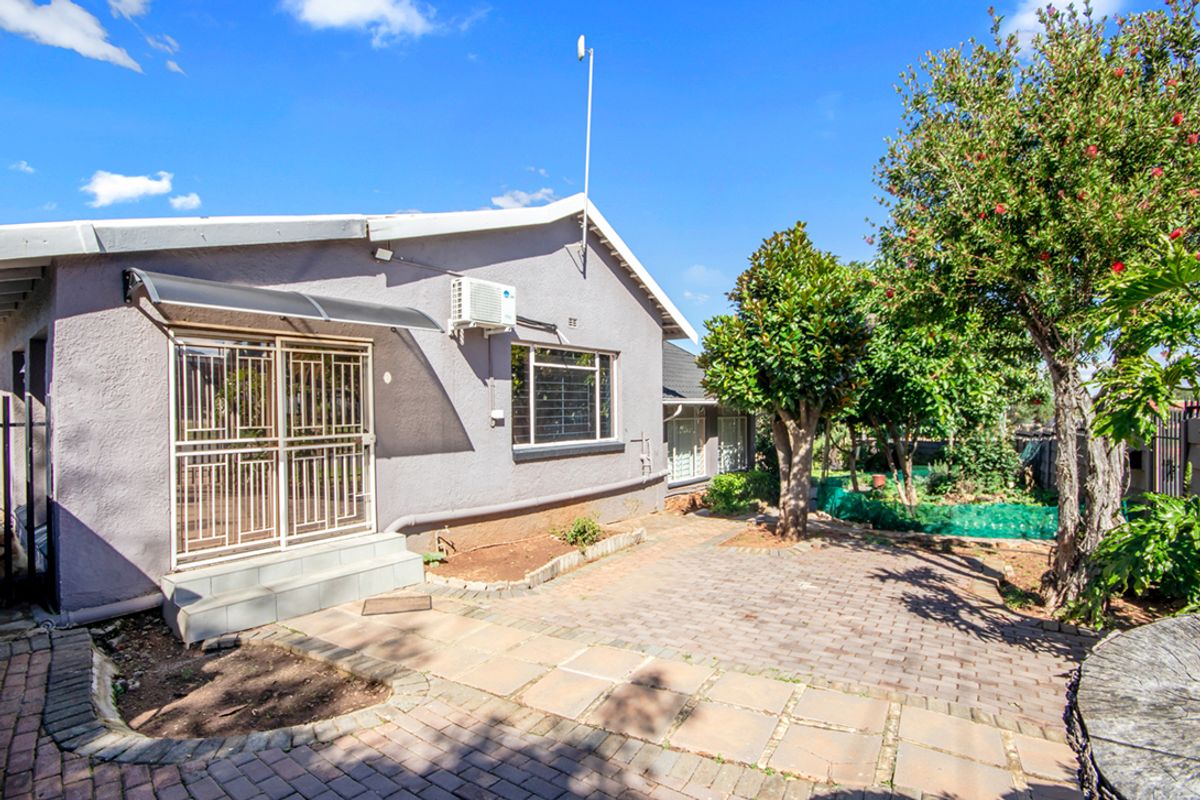 Rear exterior view of the house featuring a paved patio area, established garden trees, and a security gate entrance.