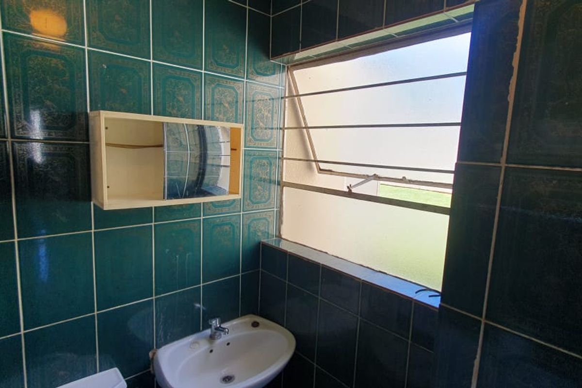 A dated bathroom featuring dark green decorative wall tiles, a wall-mounted basin, and a window with security bars.