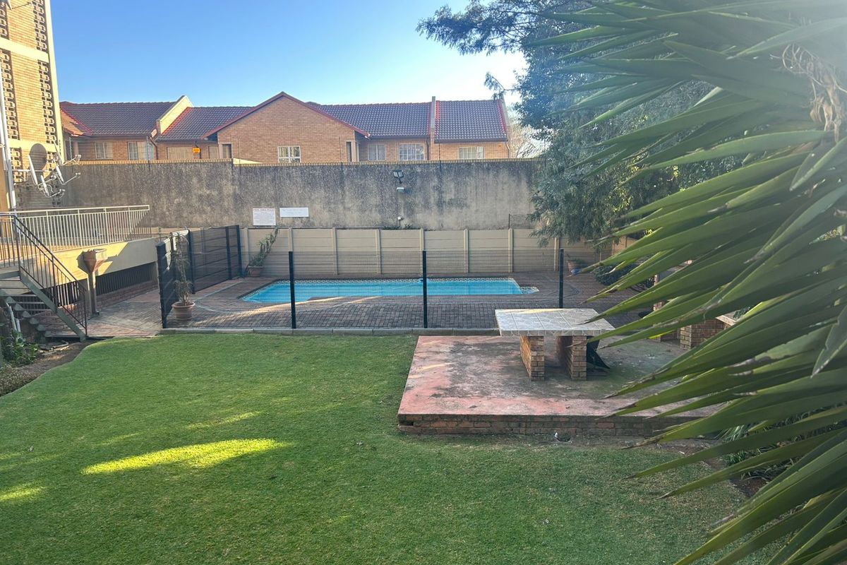 A private outdoor area featuring a swimming pool, paved patio space, built-in brick table, and a well-maintained lawn within a secure complex setting.