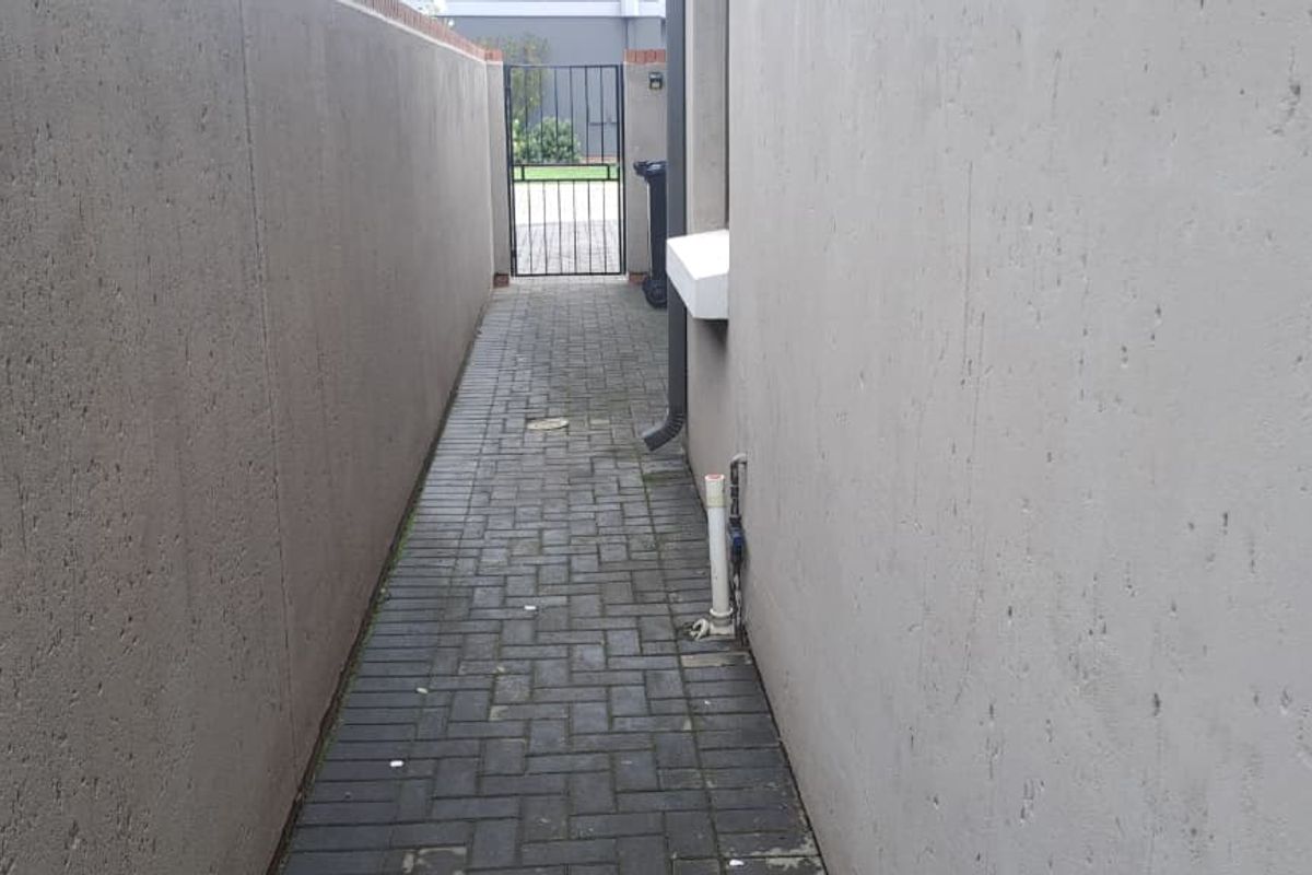 A narrow side passage featuring paved flooring, enclosed by high boundary walls, leading to a metal security gate at the rear.