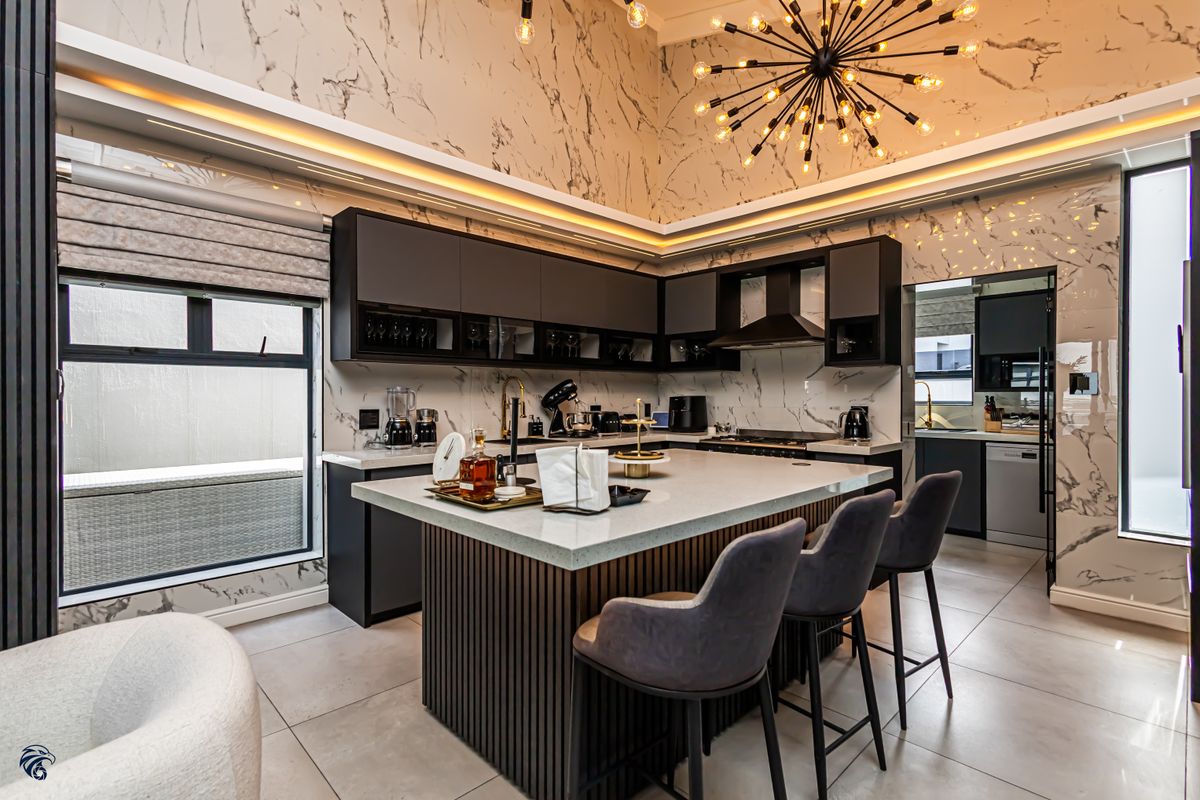 Modern luxury kitchen featuring marble-look wall cladding, a central island with stone countertop, dark cabinetry, and integrated lighting, with an adjacent scullery area.