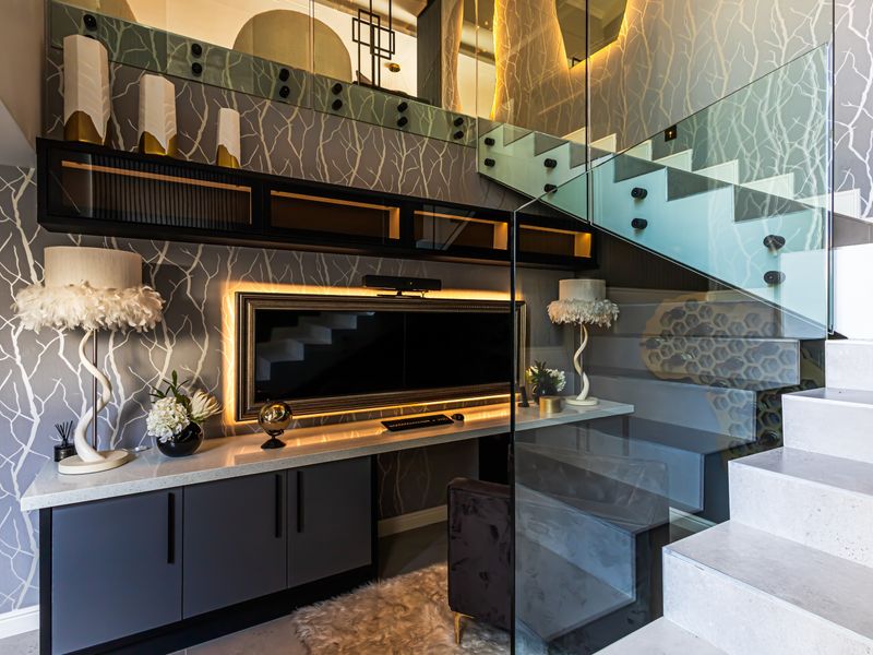 A modern, high-end home office space featuring custom built-in cabinetry, a sleek desk, and a striking glass-railed staircase with decorative wallpaper.