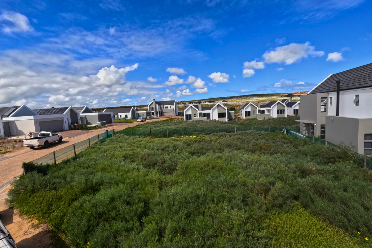 An elevated view of a vacant residential plot covered in natural vegetation, situated within a modern housing estate with surrounding contemporary homes.