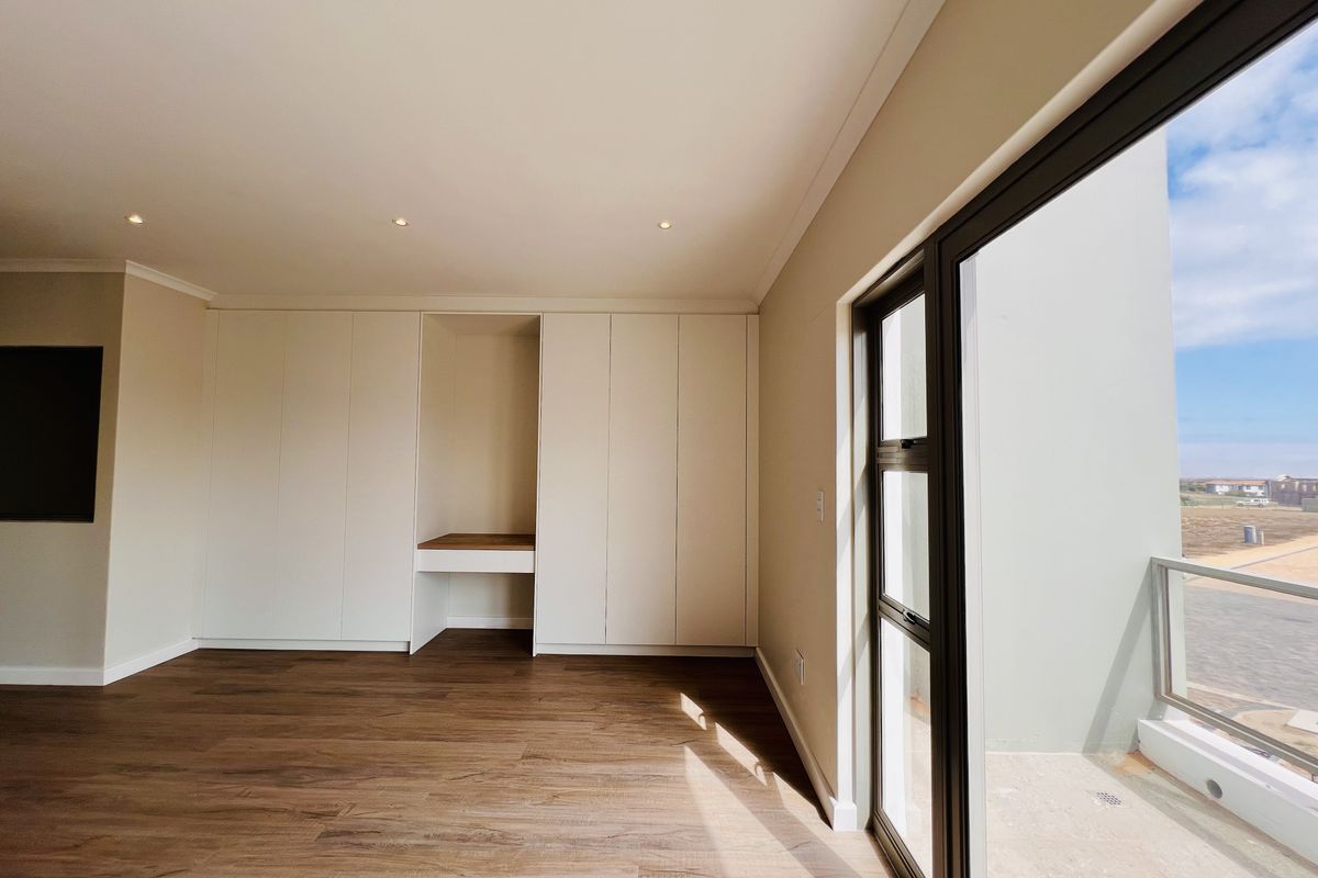A bright, modern bedroom featuring built-in cabinetry with a integrated desk space, wood-look flooring, and large glass sliding doors leading to a private balcony.