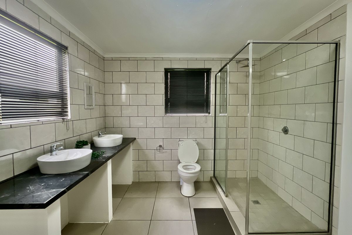 Modern bathroom featuring a double vanity with vessel sinks, a walk-in glass shower, and a toilet, finished with neutral wall and floor tiling.