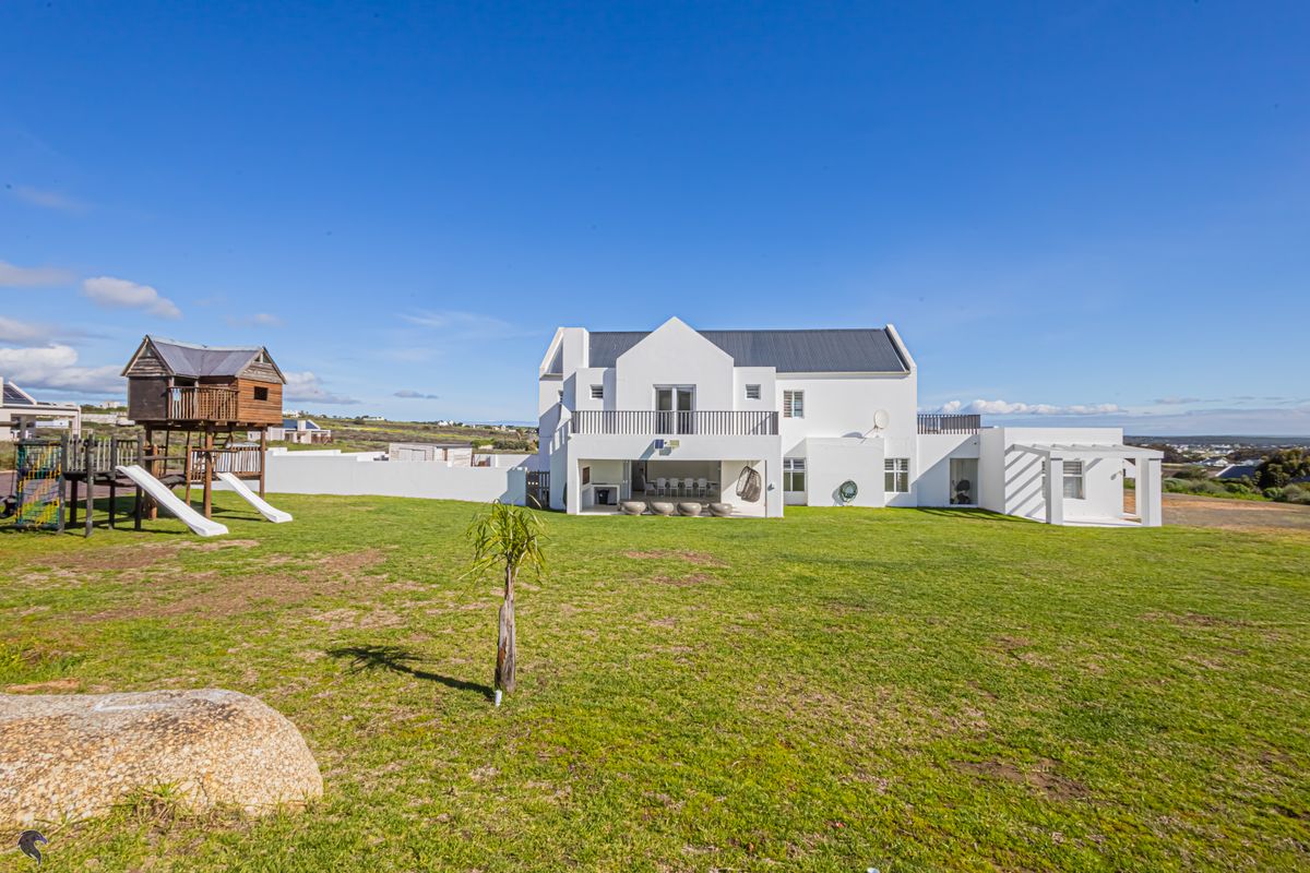 Rear view of a modern white house featuring a spacious lawn, covered patio, balcony, and a pergola, set in a large open garden.