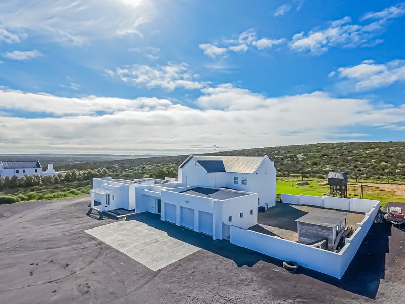 Aerial view of a white modern farmhouse with a metal roof, attached garage, and surrounding boundary wall set in a rural landscape.