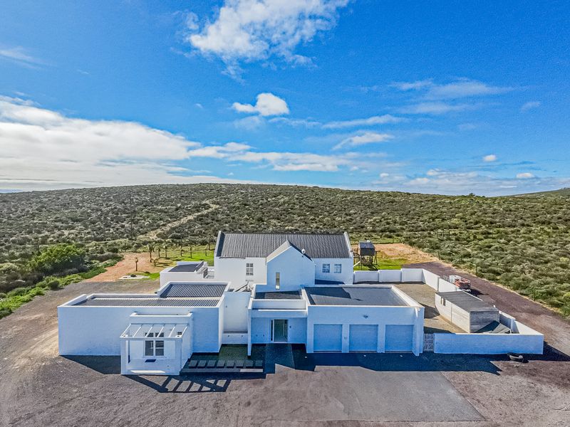Aerial view of a modern white house with a metal roof, multiple garage bays, and surrounding boundary walls set in a rural landscape.