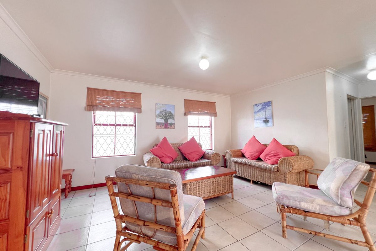A spacious living room featuring neutral floor tiles, large windows with security bars, and simple ceiling lighting.