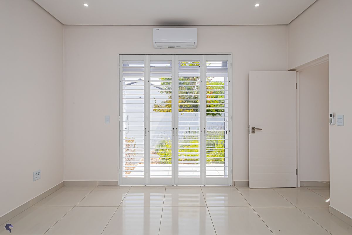 A bright, empty living space featuring tiled flooring, recessed ceiling lighting, a wall-mounted air conditioning unit, and white plantation-style shutters opening to the garden.