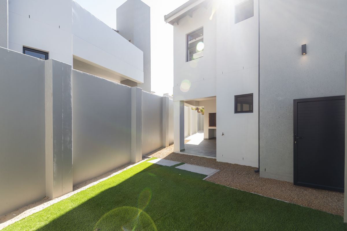 A modern rear exterior view featuring a small garden area, paved pathway, and a covered patio space with a built-in braai.