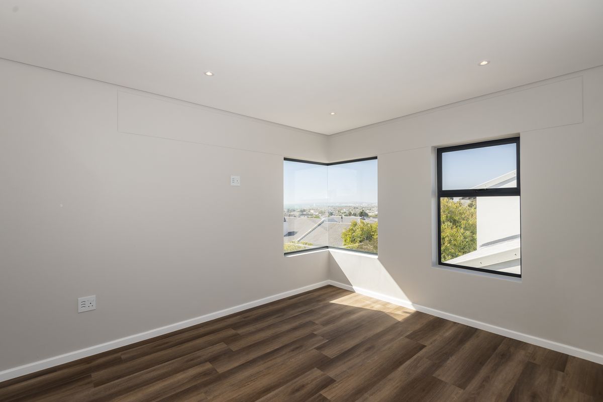 A bright, modern bedroom featuring wood-look flooring, recessed ceiling lighting, and a corner window offering expansive views.