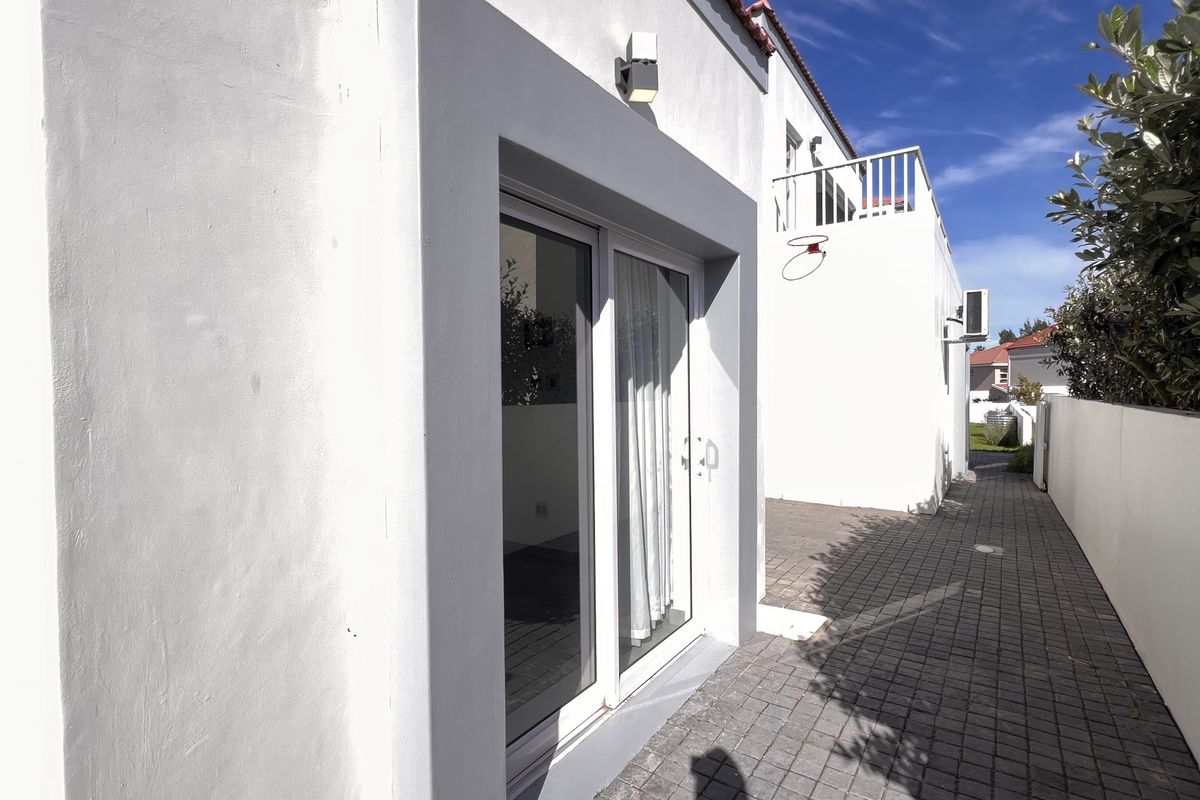 A side exterior view of the property featuring paved walkways, sliding glass doors, and an upper-level balcony with white railings.