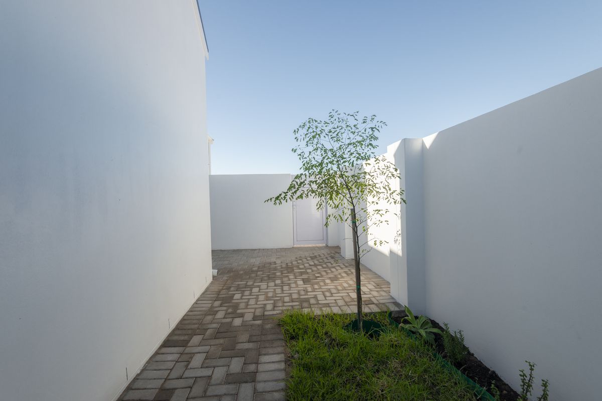 A clean, minimalist side passage or courtyard area featuring paved walkways, white boundary walls, and a small landscaped garden section.