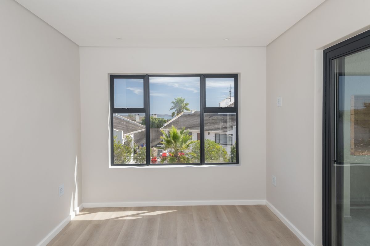 A bright, empty bedroom featuring modern light wood-look flooring, neutral walls, and large dark-framed windows offering a pleasant neighbourhood view.