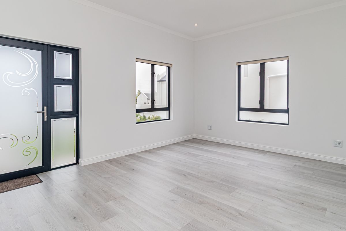 A bright, empty living room featuring modern light-toned laminate flooring, neutral walls, and dark-framed windows and entrance door.