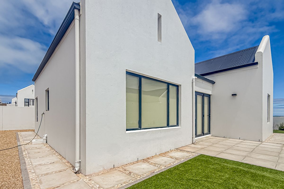 A view of the rear exterior of the house, featuring a paved patio area, gravel side path, and modern white facade with dark window frames.