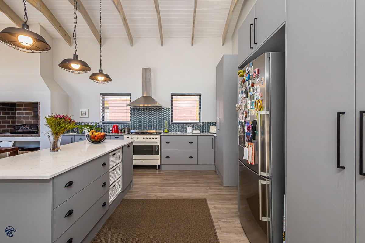 Modern kitchen featuring a central island, quartz countertops, grey cabinetry, gas stove with extractor, and exposed timber ceiling beams, adjacent to a brick fireplace.