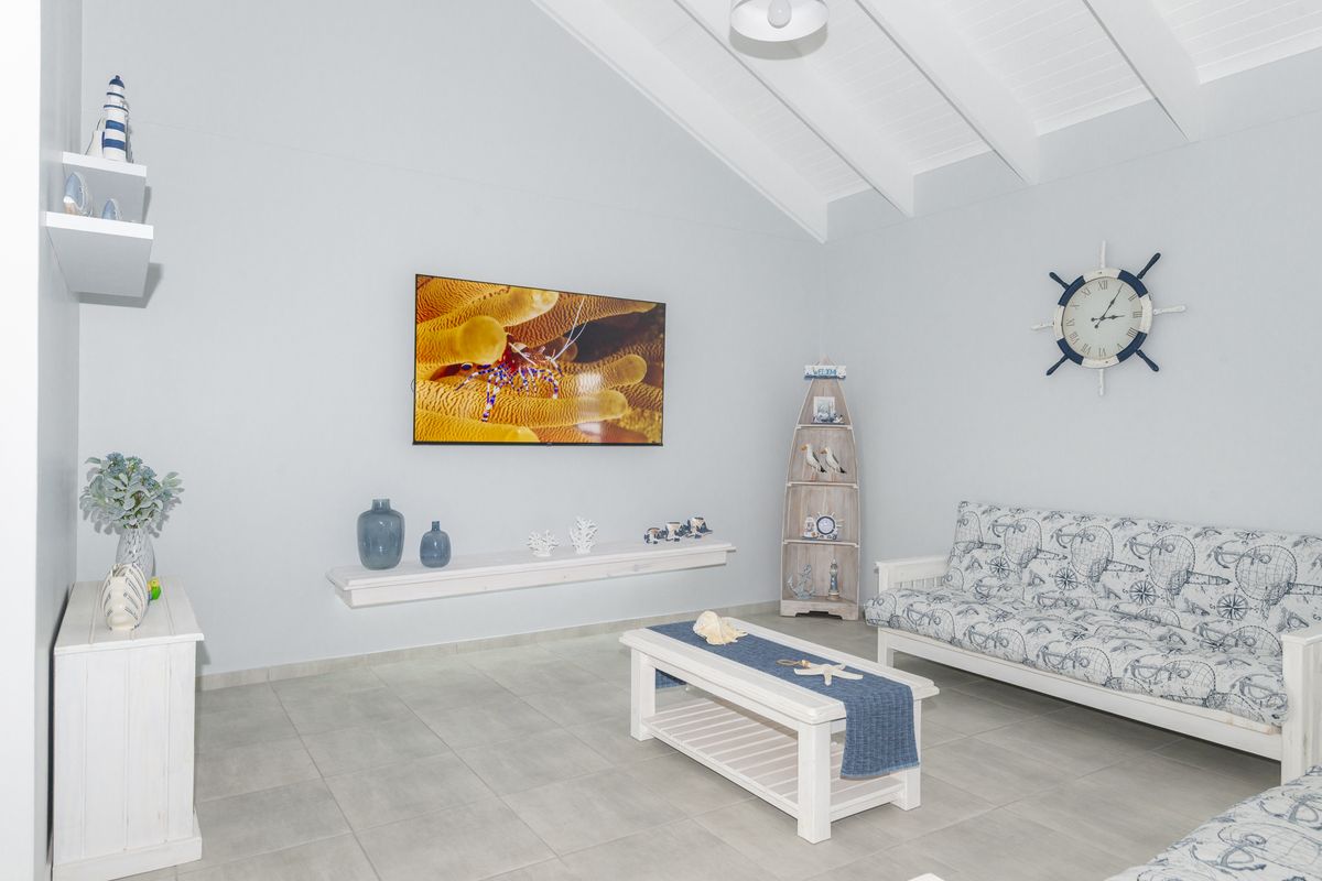 A bright living room featuring vaulted ceilings with exposed beams, neutral tiled flooring, and a wall-mounted television.