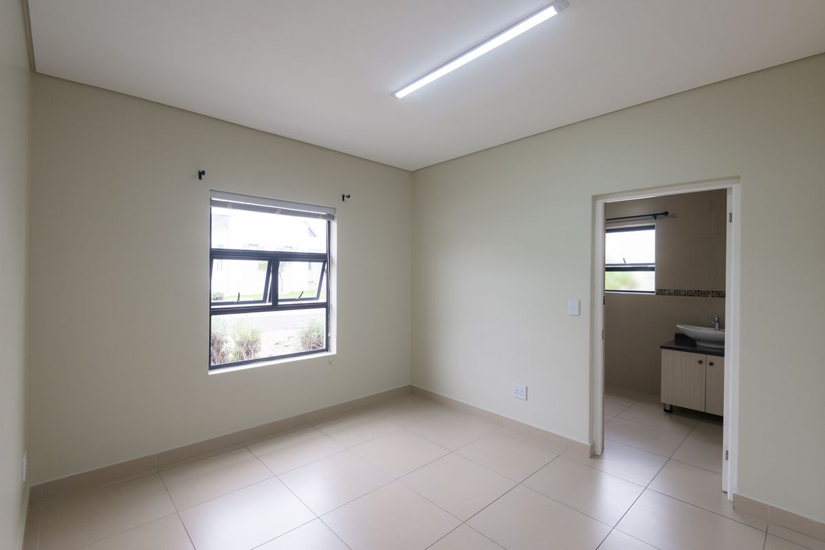 A bright, empty bedroom featuring neutral tiled flooring, an aluminium window, and direct access to an en-suite bathroom.