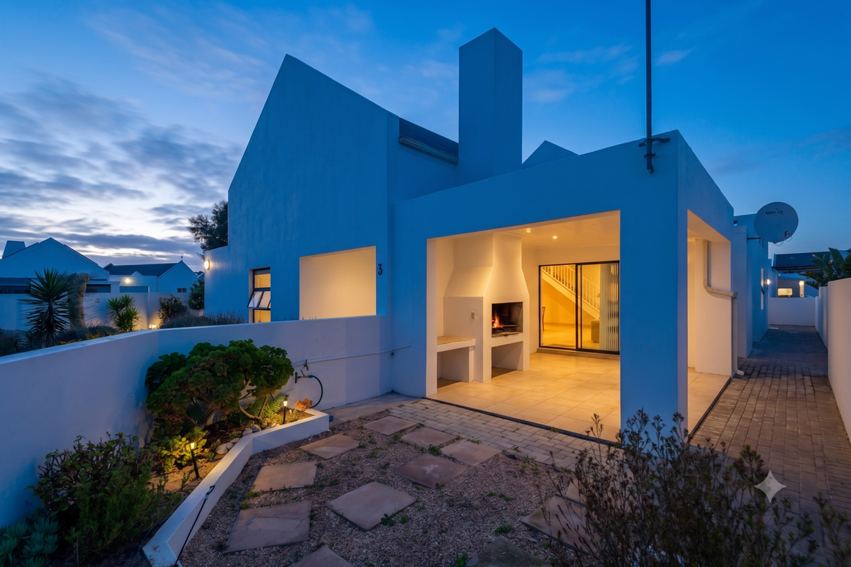Rear exterior view featuring a covered patio with a built-in braai, paved walkway, and landscaped garden area at dusk.
