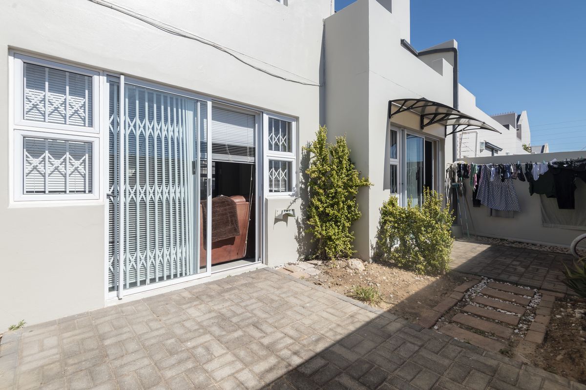 A rear exterior view featuring a paved patio area, sliding glass doors with security gates, and a small garden space.