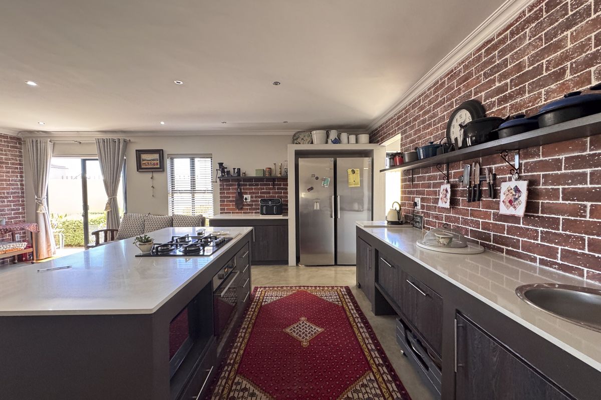 Modern open-plan kitchen featuring a large central island with integrated gas hob, stone countertops, dark cabinetry, and a distinctive exposed brick feature wall.