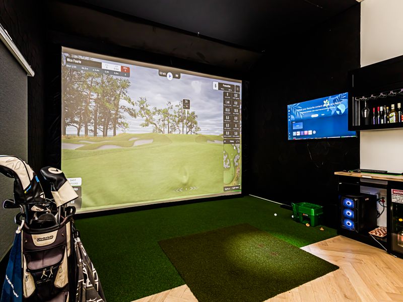 A dedicated indoor golf simulator room featuring a large projection screen, artificial turf hitting area, and integrated bar cabinetry.