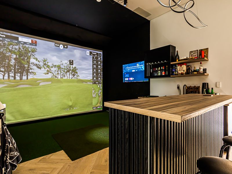 A modern home sports facility featuring a professional golf simulator setup, a custom built-in bar with shelving, and stylish herringbone flooring.