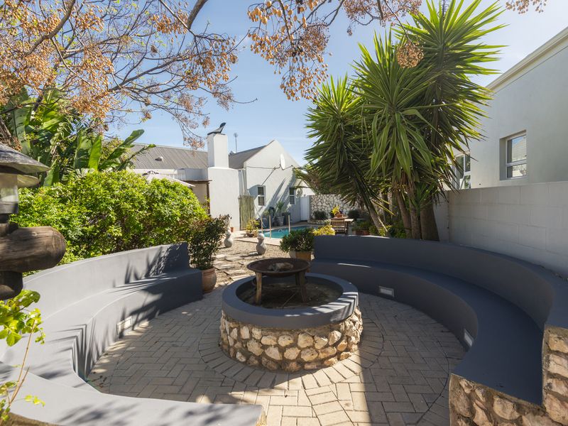 A well-maintained rear garden featuring a paved patio area with a built-in fire pit, curved masonry seating, and a swimming pool in the background.