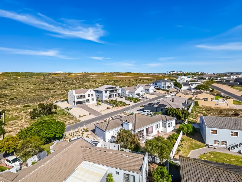 Aerial view of a coastal residential neighbourhood featuring modern homes with tiled roofs, paved driveways, and solar installations under a clear blue sky.